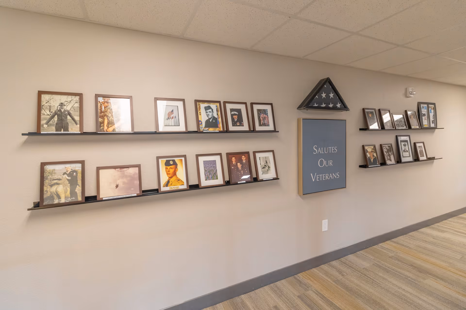 A hallway wall display featuring framed photographs of veterans and a folded American flag in a triangular case. A sign on the wall reads 'Salutes Our Veterans'. The floor has a striped carpet and the ceiling has white tiles.