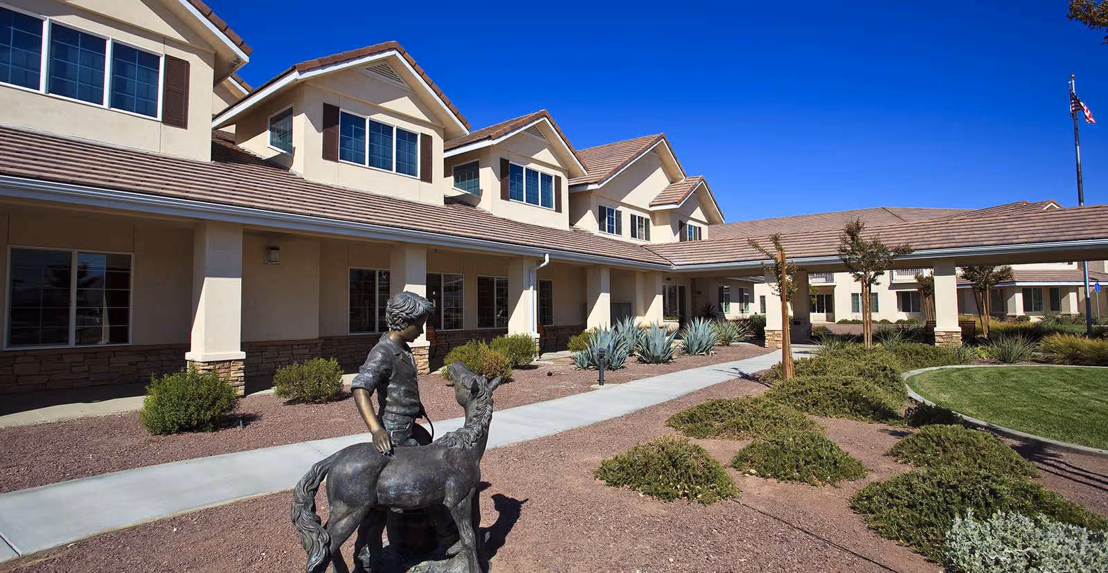 Exterior view of a senior living facility building with beige walls and brown roof tiles under a clear blue sky. In the foreground, there is a bronze statue of a child with a horse. The landscaping includes bushes, small trees, and a curved walkway leading to the entrance.