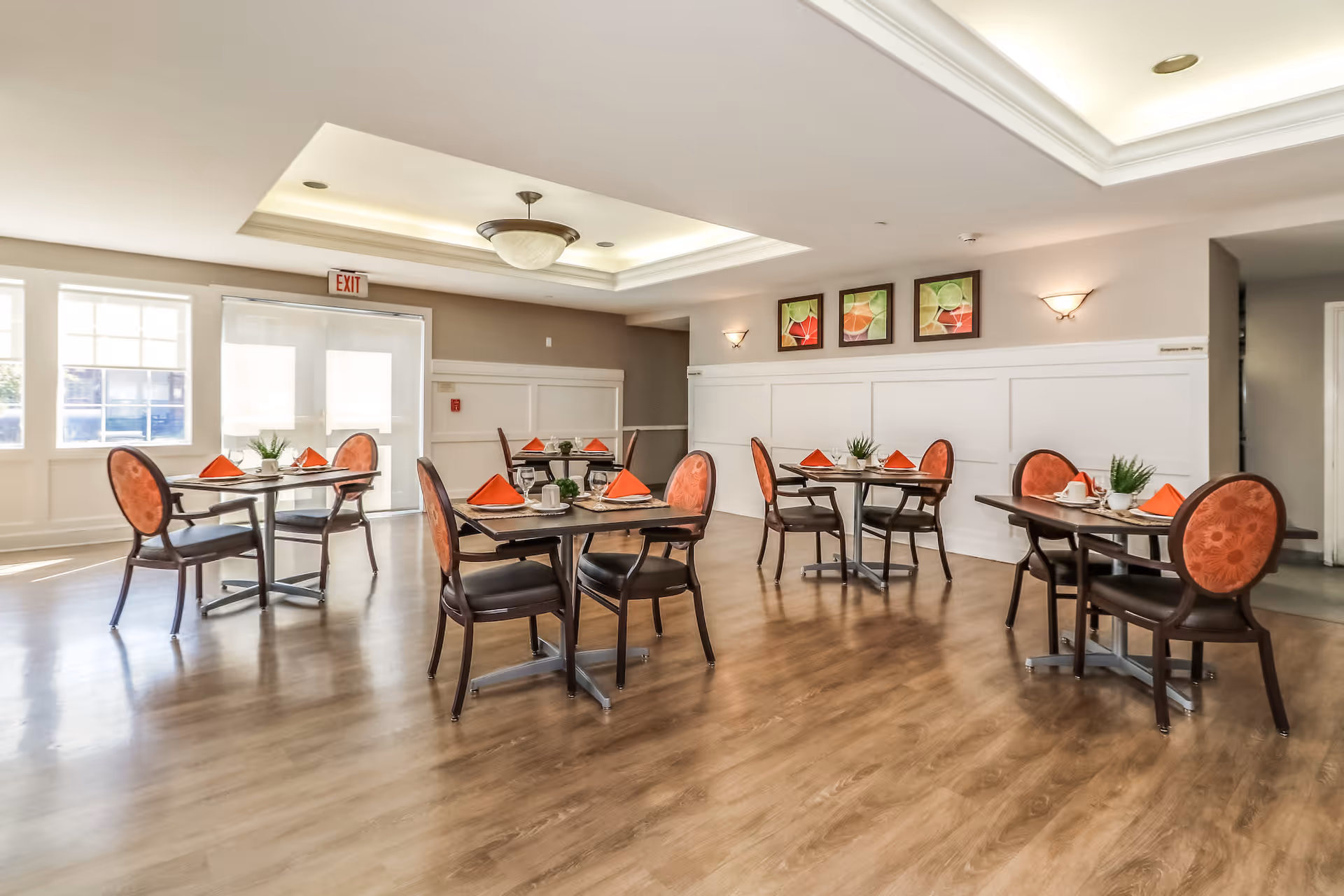 A bright dining room with wooden floors and several small round tables, each set with orange napkins, white cups, and small green plants. The room has large windows letting in natural light, a ceiling light fixture, and three colorful framed artworks on the wall.