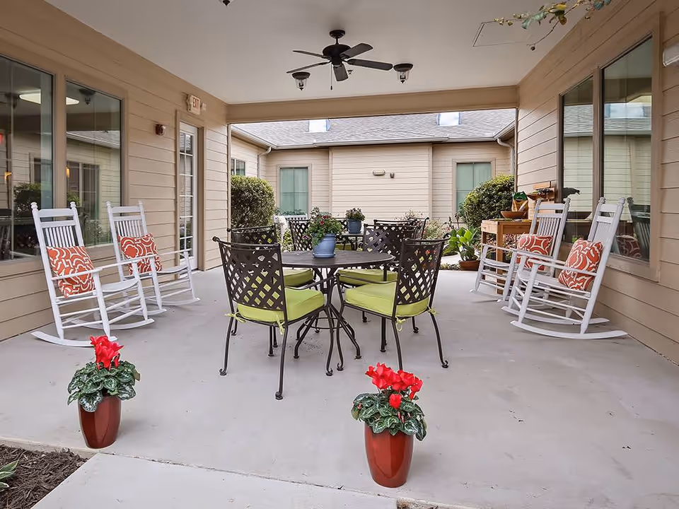 Covered courtyard patio with a round table and metal chairs, white rocking chairs with patterned cushions, and potted plants.