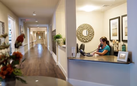 Reception area inside Vantage Pointe Village with a woman sitting at the front desk talking on the phone. The desk has a computer, phone, and decorative items. A long hallway with wooden flooring and several doors extends into the background. The walls are light-colored with framed artwork and a decorative round mirror behind the desk.