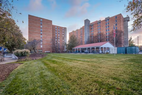 Exterior view of a multi-story brick building with a large green lawn in the foreground, a covered patio area with a red roof, and an American flag on a flagpole. Trees and shrubs surround the building under a partly cloudy sky.