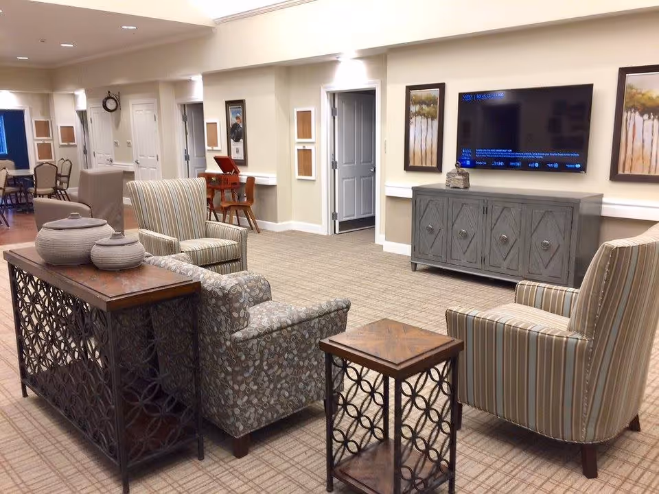A cozy living room area in a senior living facility featuring patterned armchairs and a small wooden side table. A large flat-screen TV is mounted on the wall above a gray wooden cabinet. The room has beige walls with framed artwork and a carpeted floor. In the background, there is a small desk with a chair and additional seating visible.