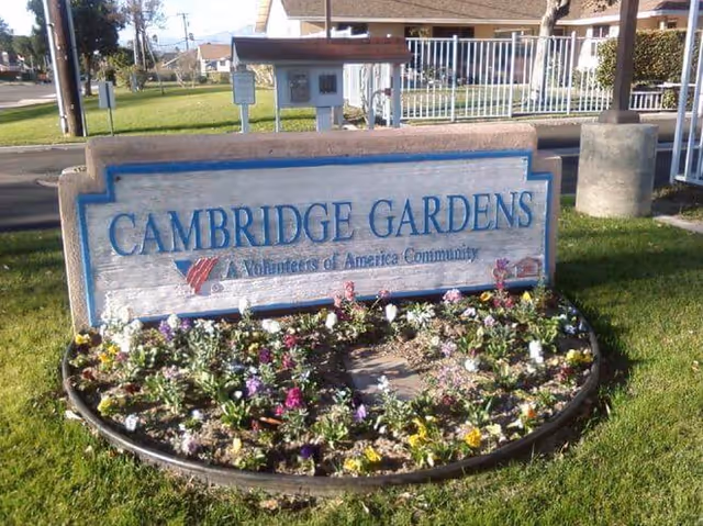 A Cambridge Gardens sign mounted on a low wall, surrounded by a circular flower bed and a manicured lawn with a white fence in the background.