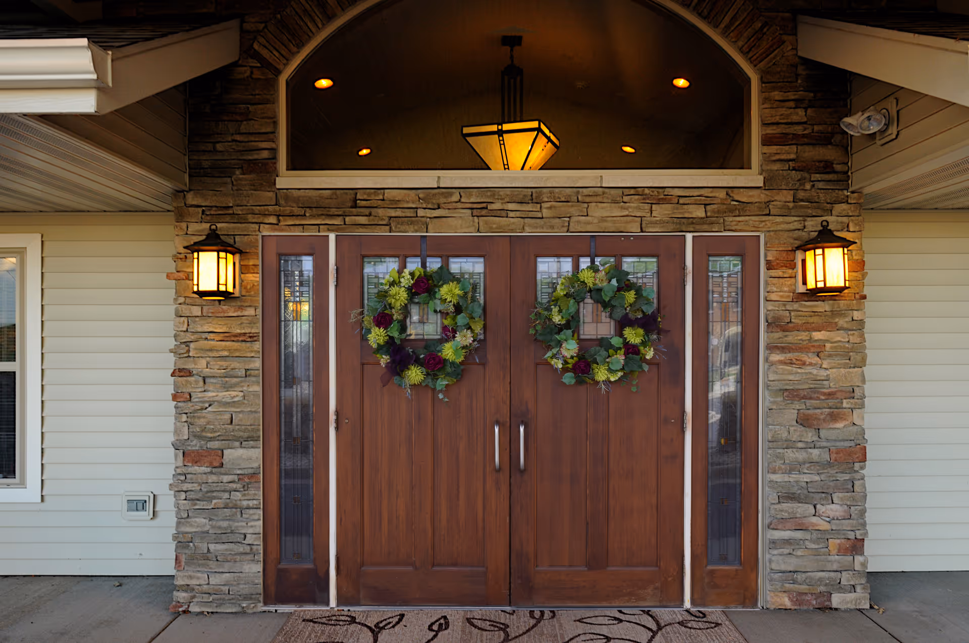 Double wooden entrance doors with wreaths set in a stone façade flanked by lanterns and a transom window above.
