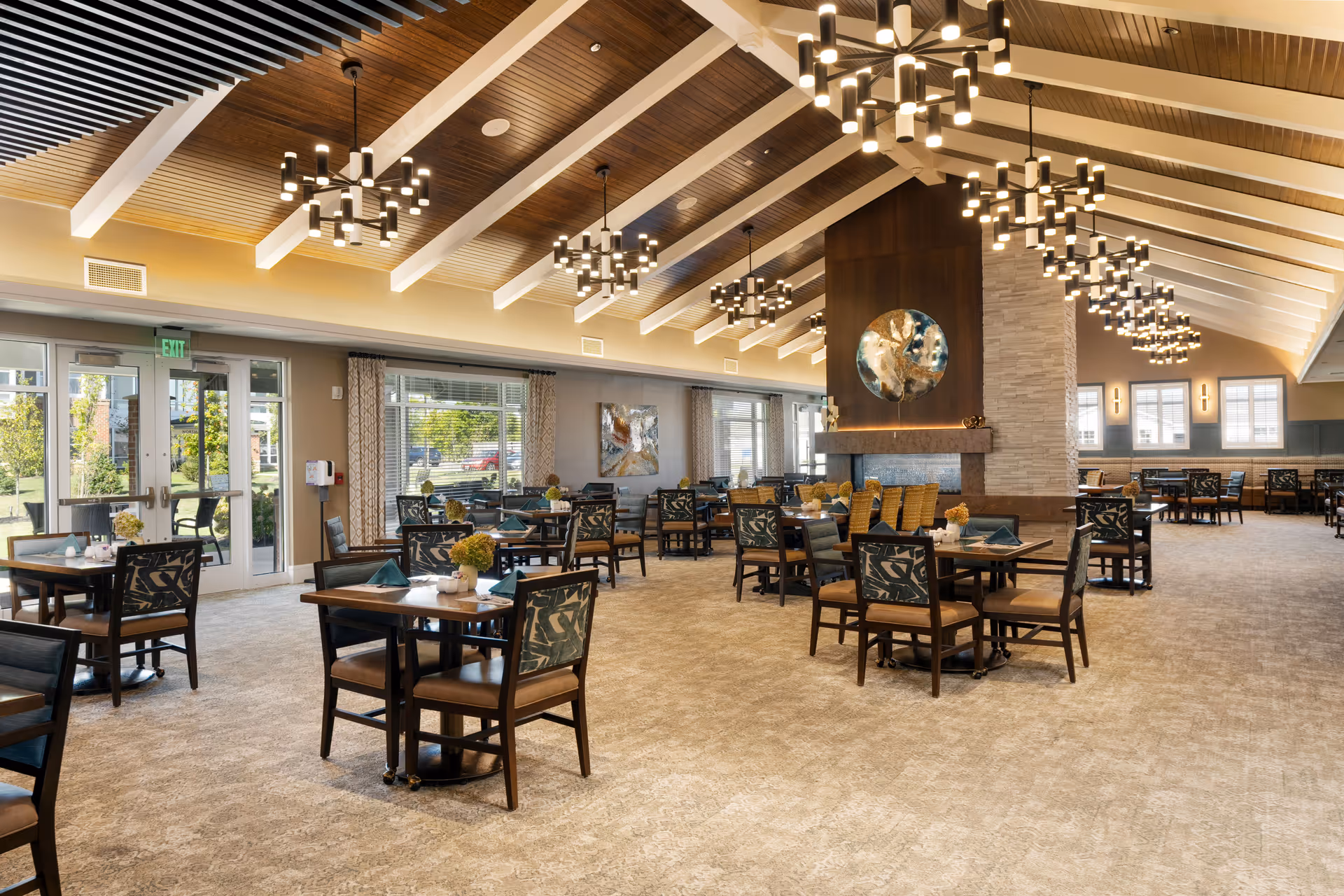 Spacious dining room with multiple wooden tables and chairs arranged neatly. The room features a high ceiling with wooden beams and modern chandeliers. Large windows and glass doors allow natural light to fill the space. A central fireplace with decorative art above it divides the room.