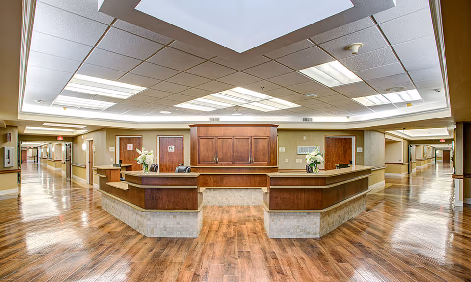 Spacious nurses' station and reception desk in a well-lit senior living facility hallway with wood floors and ceiling lights.