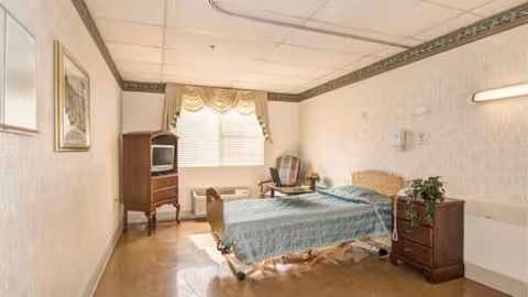 A furnished bedroom in a senior facility featuring a hospital bed, dresser, and window.