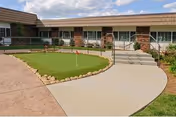 Outdoor courtyard area at a senior living facility featuring a small putting green with a red flag, surrounded by a concrete walkway and landscaping with rocks and plants. The building exterior has large windows and a covered walkway with brick pillars.