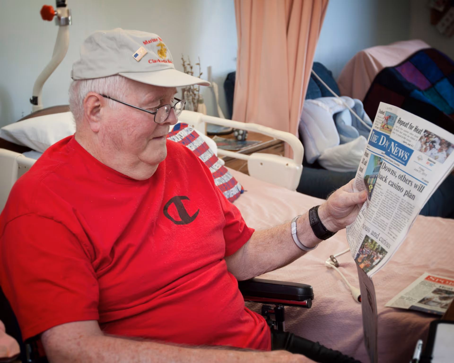 An elderly man wearing a red shirt and a beige cap is sitting in a wheelchair inside a room, reading a newspaper. Behind him is a bed with a pillow and a blanket, and some furniture including a chair and a table with items on it.