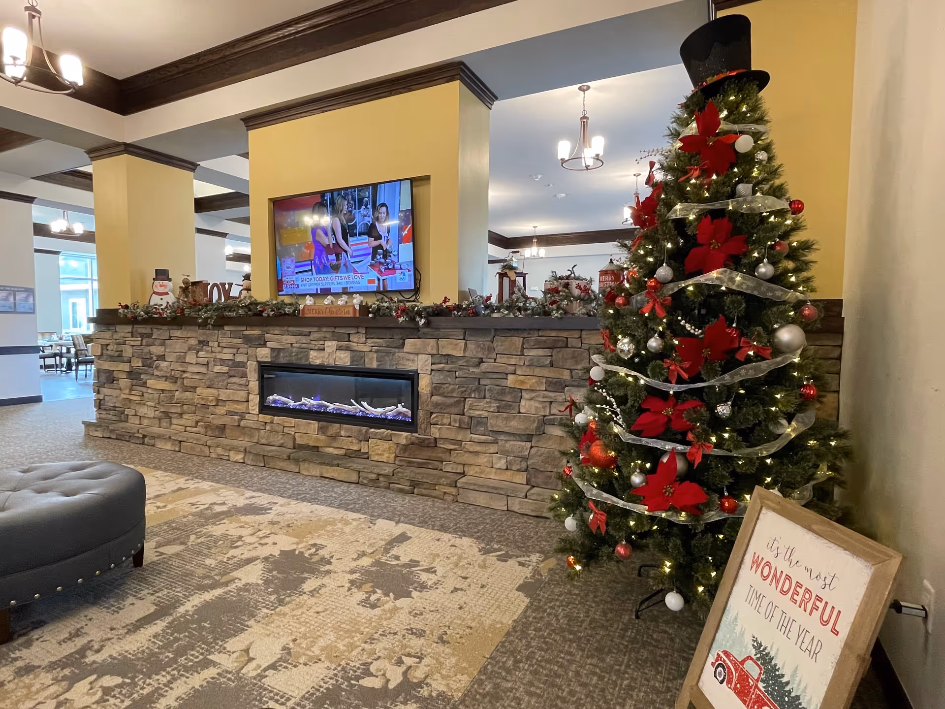 Interior view of a senior living facility lobby decorated for Christmas with a Christmas tree adorned with red poinsettias, silver ribbons, and ornaments. A stone fireplace with a mounted TV above it is visible, along with festive garlands and holiday decorations. A sign next to the tree reads 'It's the most wonderful time of the year.' The area has carpeted flooring, beige walls, and dark wood trim.