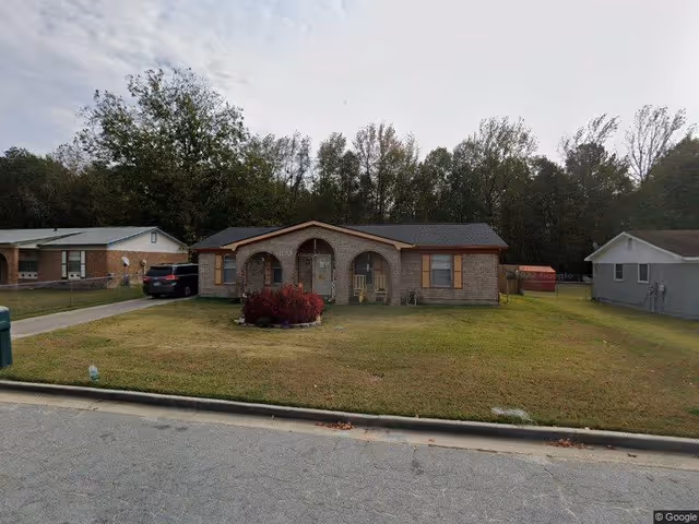 Single-story brick house with a front porch featuring two arches, a small garden with red bushes in the front yard, and a driveway with a black vehicle parked. The house is surrounded by grass and trees in the background under a cloudy sky.