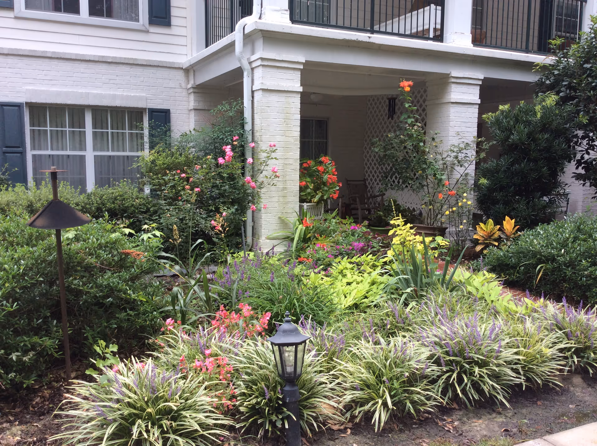 Landscaped front garden with flowering plants and a covered ground-floor porch of a light-brick building.