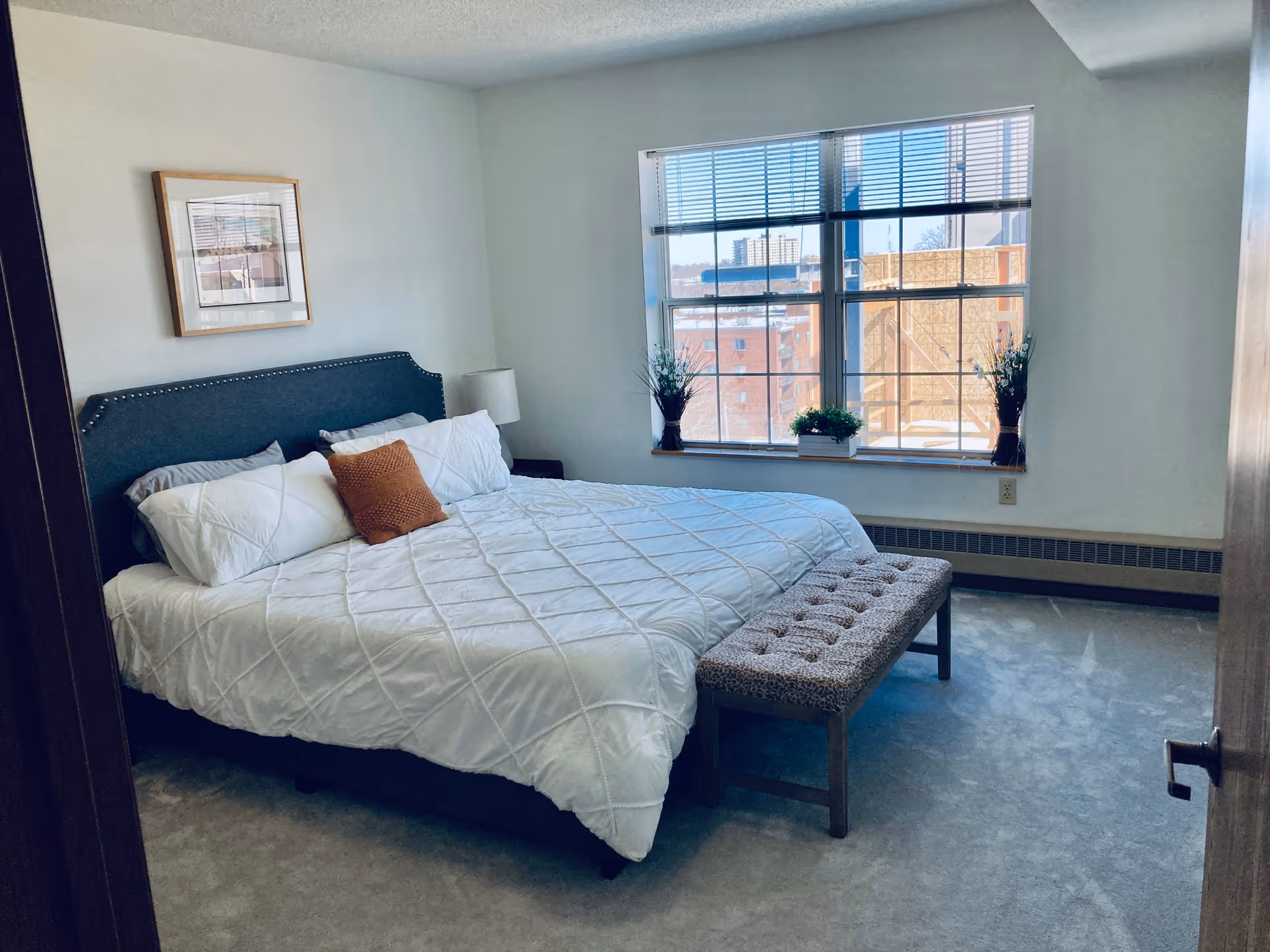 Bright bedroom with a made bed, tufted bench at the foot, bedside lamp, and a large window with plants overlooking neighboring buildings.