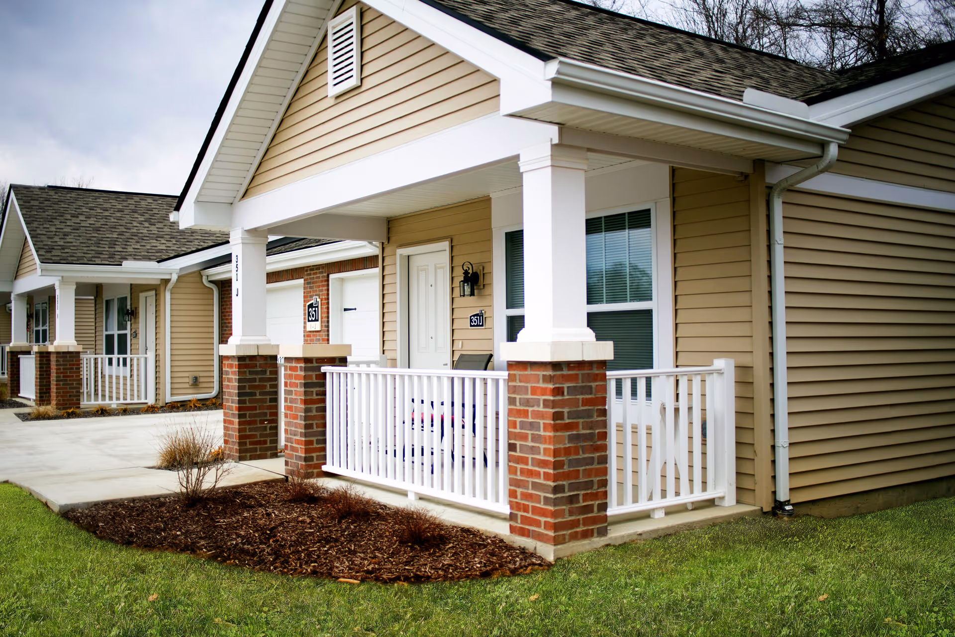 Row of single-story beige residential units with covered porches, white railings, brick columns, and small lawns.