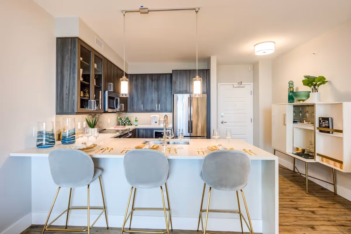 Modern kitchen with dark wood cabinets, stainless steel refrigerator and microwave, and a white island countertop with three gray bar stools. The island is set with plates, glasses, and gold-colored cutlery. Pendant lights hang above the island, and there is a white cabinet with decorative items on the right side of the room.