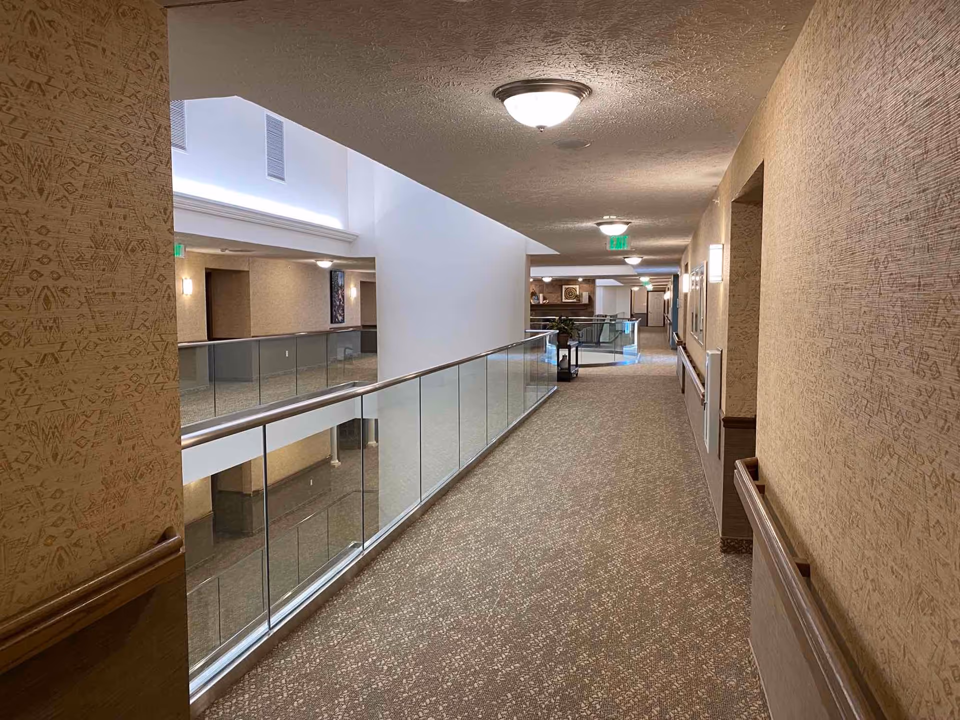 Upper-level interior hallway in a retirement community with carpeted floor, glass railing overlooking an atrium, and ceiling lights.