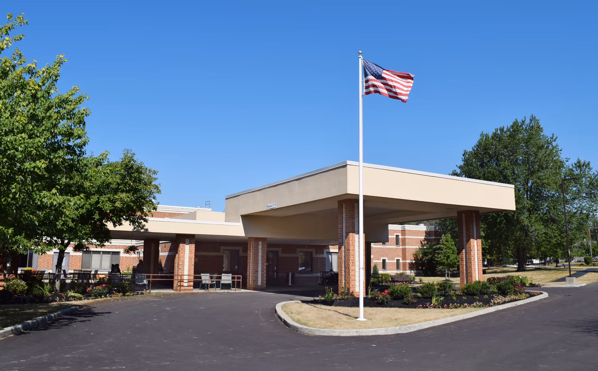Exterior view of Elderwood Assisted Living at Cheektowaga showing the entrance with a covered drop-off area supported by brick columns, an American flag on a flagpole in front, and surrounding trees and landscaping under a clear blue sky.