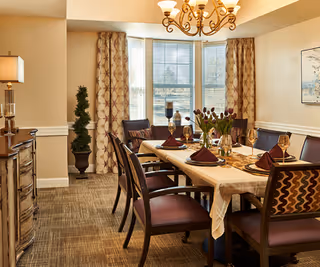 A dining room with a rectangular table set for six people with folded napkins, wine glasses, and a vase of flowers. The room has patterned curtains on the windows, a chandelier overhead, a sideboard with a lamp, and a potted plant in the corner.