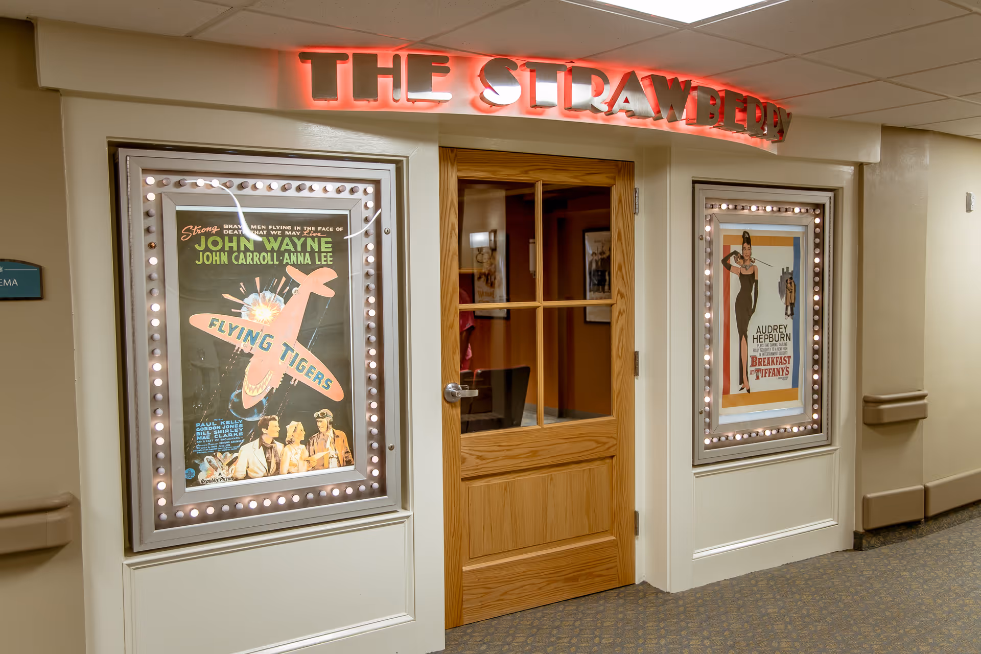 Hallway entrance to a small theater labeled "THE STRAWBERRY" with a wooden door flanked by illuminated vintage movie posters.