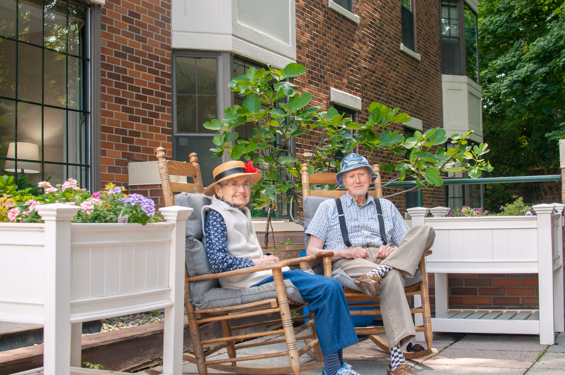 Two elderly individuals sitting on wooden rocking chairs on a patio outside a brick building. They are surrounded by white planters filled with colorful flowers and greenery. Both are wearing hats and smiling, enjoying the outdoor setting.