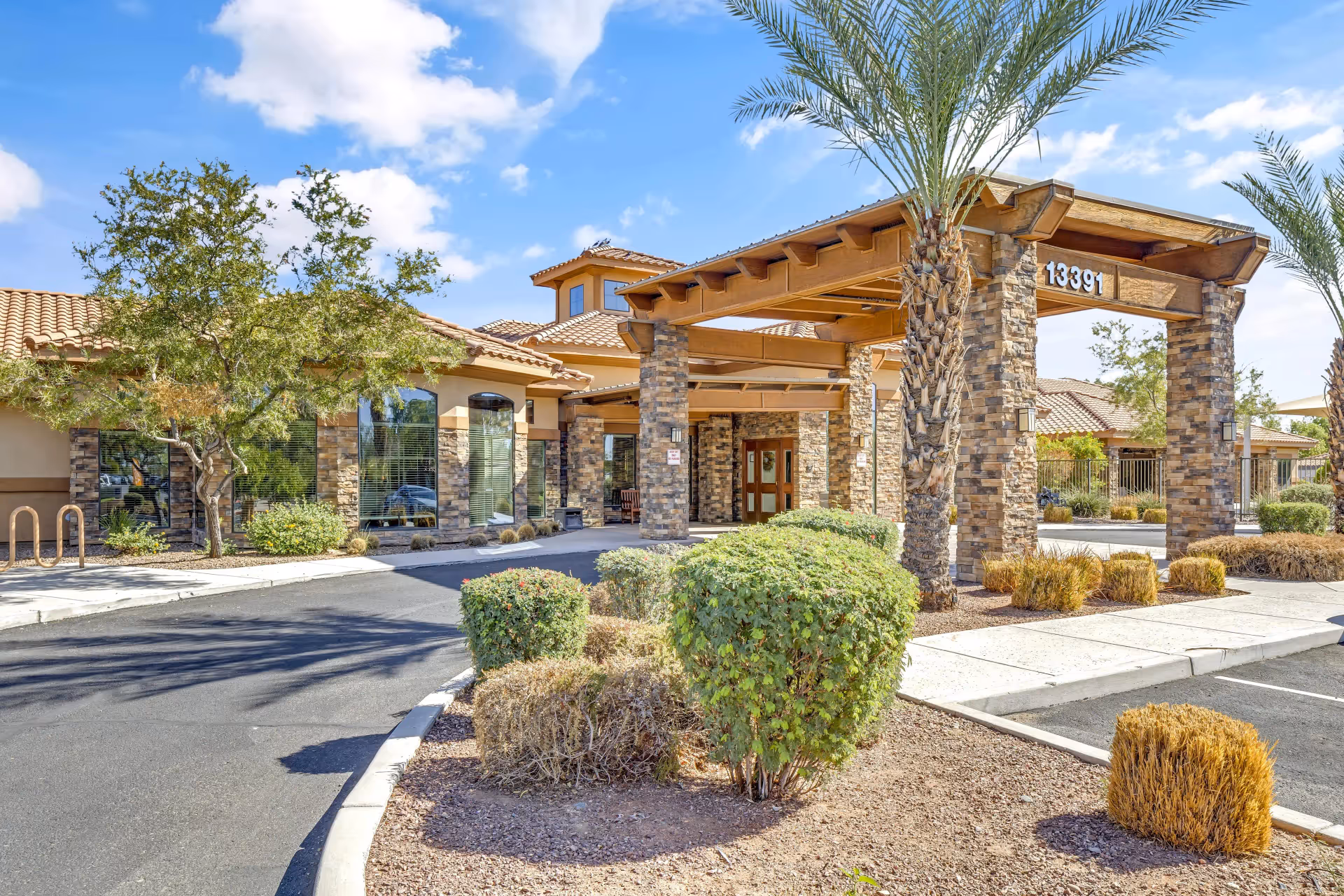 Front entrance of a senior living facility with a covered porte-cochère, stone columns, palm trees, and landscaped parking area.