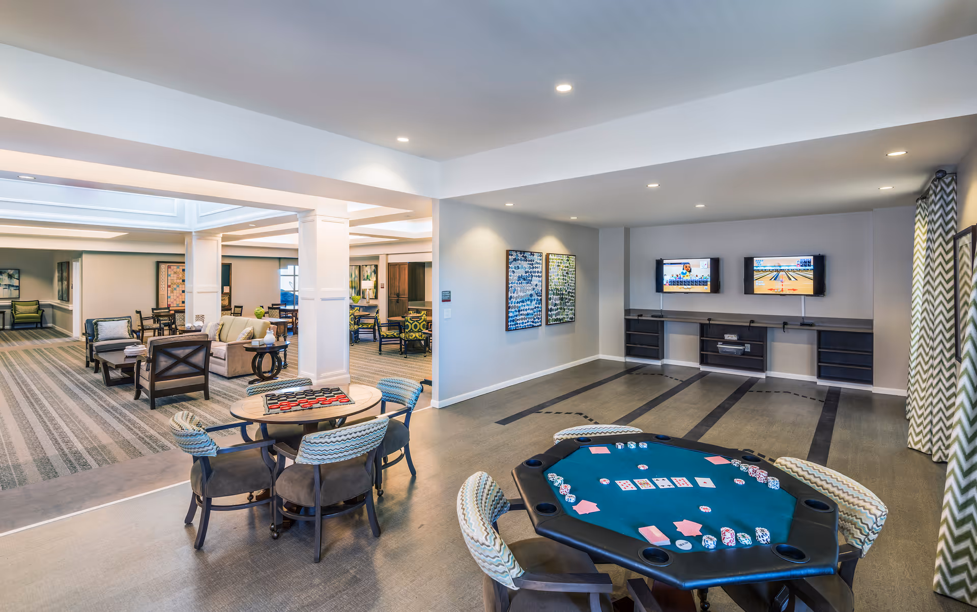 A spacious and well-lit common area in a senior living facility featuring a poker table with cards and chips in the foreground, a round table with a checkers game, multiple seating areas with chairs and sofas, two wall-mounted TVs displaying bowling games, and modern decor with patterned curtains and artwork on the walls.