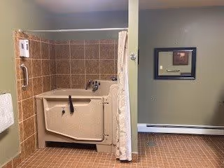 Accessible walk-in bathtub with tiled surround and shower curtain in a bathroom with green walls, towel rack, and a framed mirror.