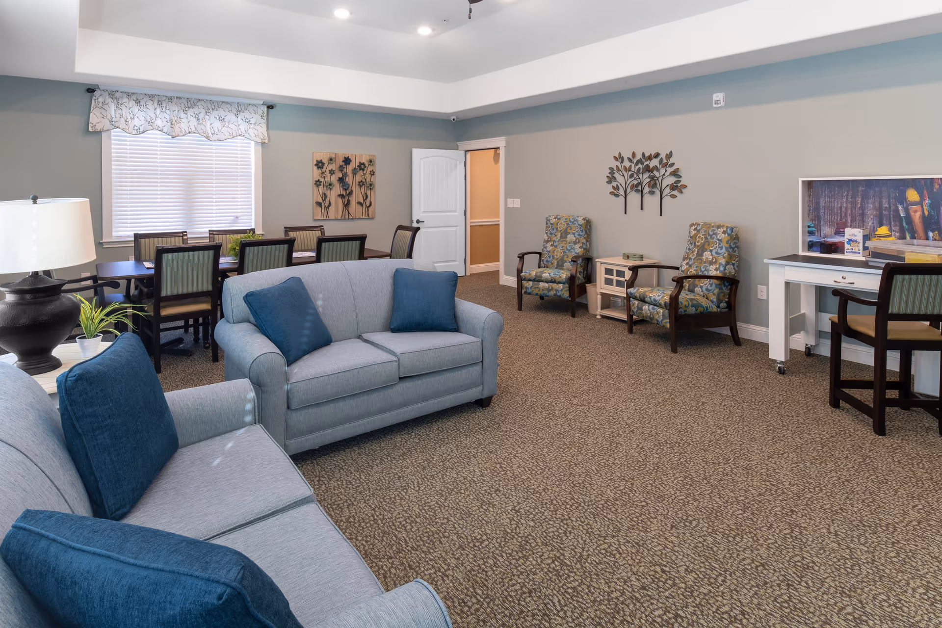 A spacious living room area in an assisted living facility featuring two light gray sofas with blue cushions, a table lamp on a side table, a dining table with chairs near a window with blinds and a floral valance, two patterned armchairs with a small table between them, and a white desk with a chair. The walls are decorated with metal wall art and a framed picture.