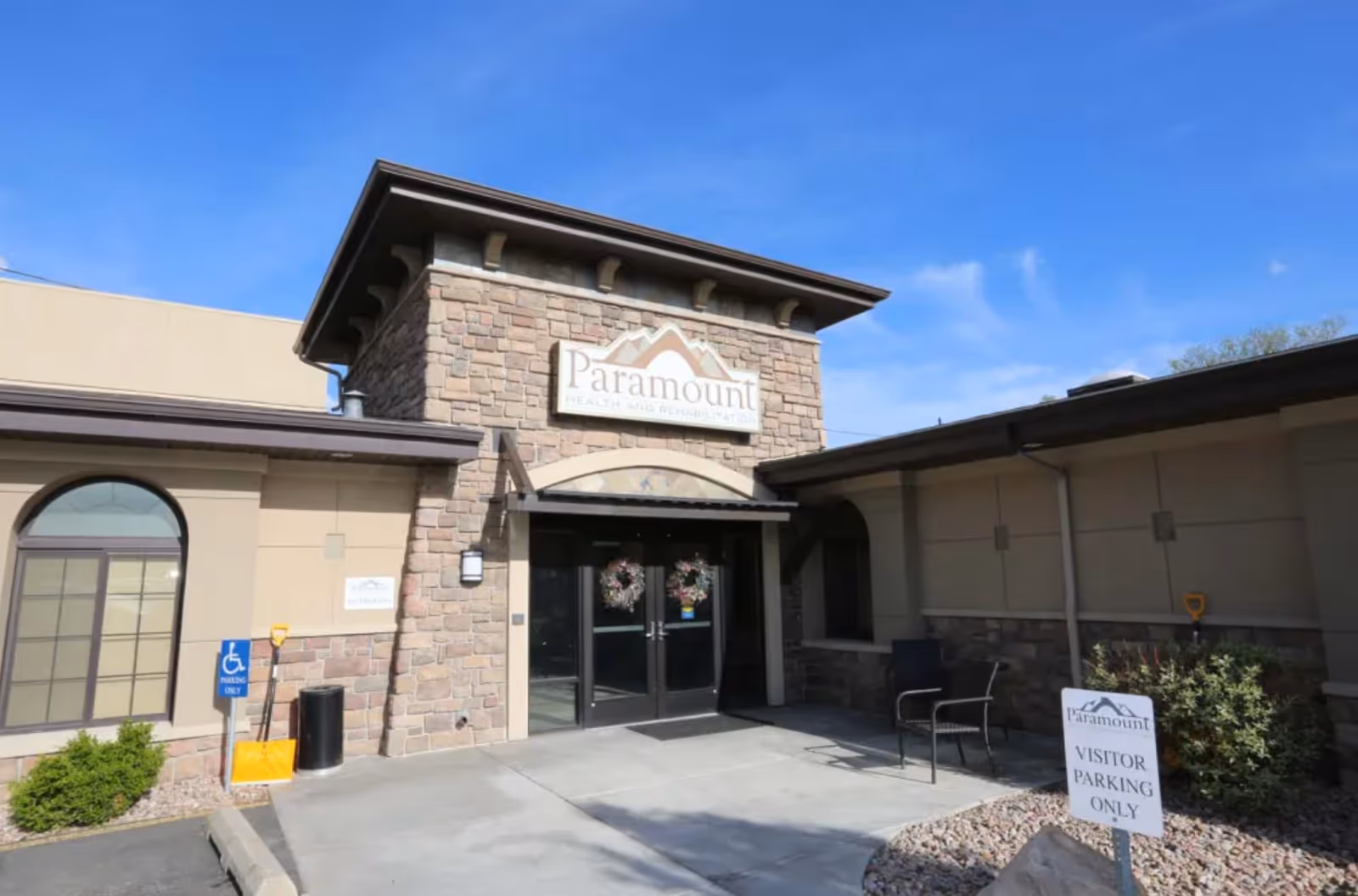 Entrance of Paramount Health and Rehabilitation facility with stone and beige exterior walls, double glass doors decorated with wreaths, a visitor parking sign, and a clear blue sky.