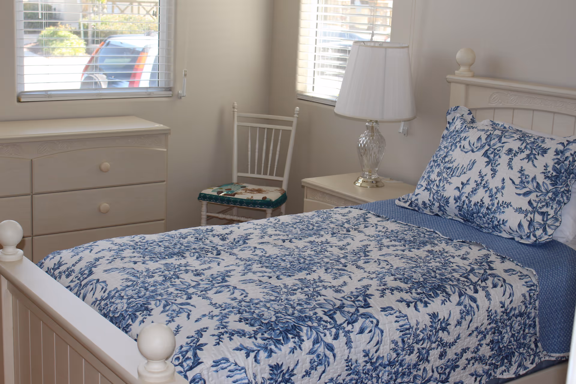 A bedroom with a white wooden bed featuring a blue and white floral patterned bedspread and matching pillow. Next to the bed is a white nightstand with a clear glass lamp with a white lampshade. There is a white wooden chair with a cushion and a white dresser under two windows with blinds partially open, letting in natural light.