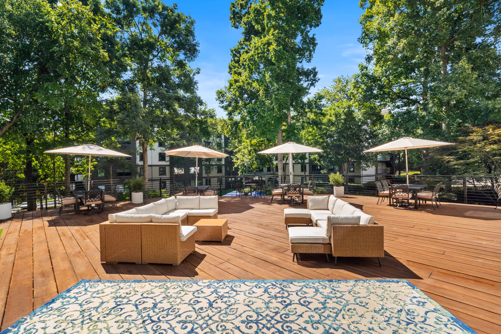 Spacious outdoor wooden deck with cushioned wicker seating arranged in a U-shape, several tables with umbrellas, surrounded by tall green trees and residential buildings in the background under a clear blue sky.