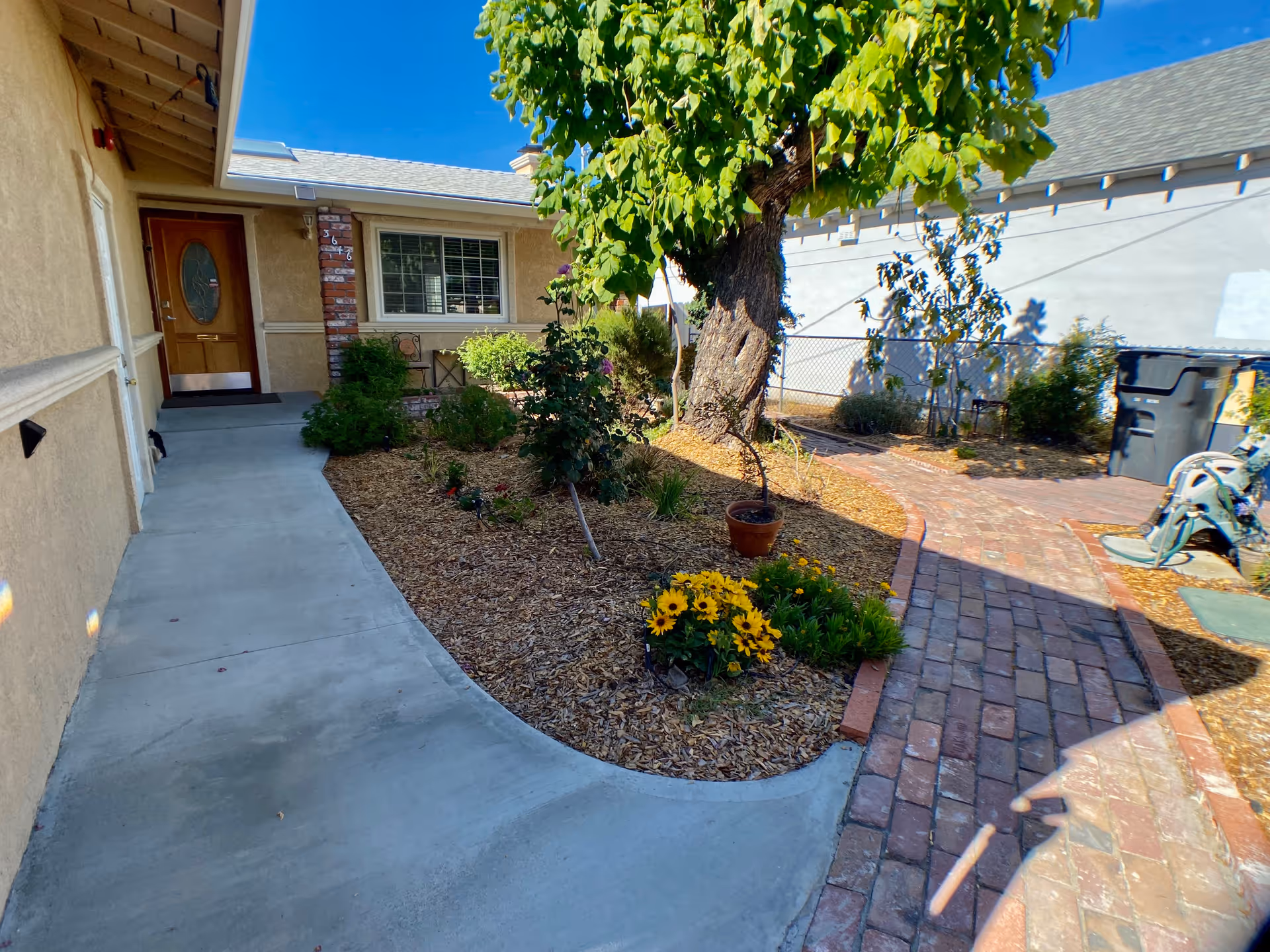 Outdoor view of a senior living facility showing a concrete walkway leading to a wooden door with an oval glass window. There is a garden area with mulch, green bushes, a tree, and yellow flowers. A brick pathway curves around the garden, and a white building wall with a window is visible in the background under a clear blue sky.