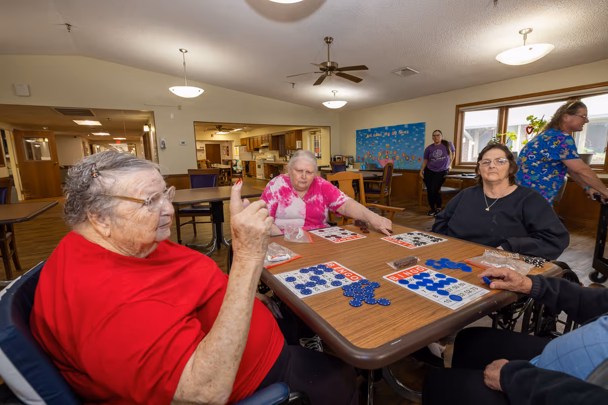 Four elderly individuals sitting around a table playing bingo in a well-lit room with wooden floors and furniture. Two women are seated on the left and right sides of the table, one wearing a red shirt and the other a black shirt. Another woman in a pink and white tie-dye shirt is seated at the far side of the table. A person in a blue patterned shirt is walking in the background near windows with plants on the sill. The room has ceiling fans and light fixtures, and a bulletin board is visible on the back wall.