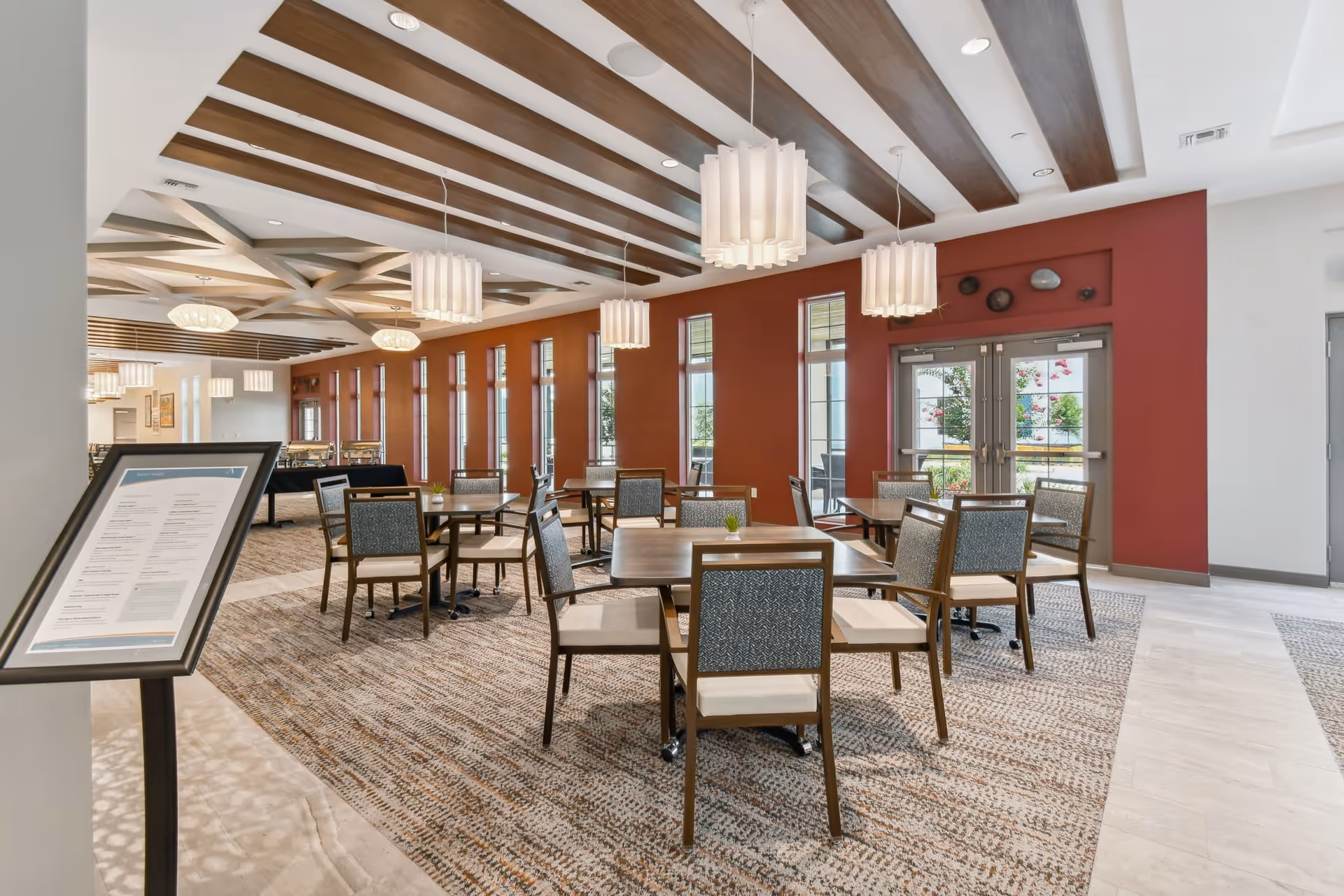 A spacious dining room in a senior living facility with multiple square wooden tables and cushioned chairs arranged neatly on a patterned carpet. The room features large windows along one wall, allowing natural light to brighten the space. The ceiling has decorative wooden beams and modern pendant lights hanging above the tables. A menu stand is visible in the foreground, and double glass doors lead to an outdoor area with greenery.