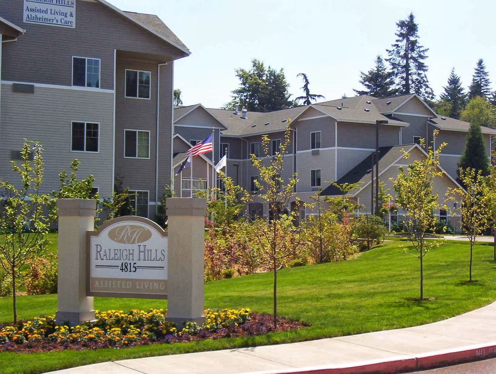 Exterior view of Raleigh Hills Assisted Living & Memory Care facility showing a multi-story building with beige and brown siding, surrounded by green lawns, small trees, and flower beds. A sign in the foreground reads 'Raleigh Hills 4815 Assisted Living'. American and other flags are visible near the building.