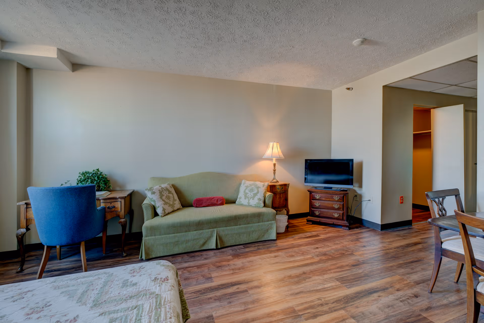 A cozy living room area featuring a green sofa with decorative pillows and a red bolster pillow, a wooden side table with a lamp, a small flat-screen TV on a wooden dresser, a blue upholstered chair at a wooden desk with a plant, and a wooden dining table with chairs. The room has wood flooring and neutral-colored walls.