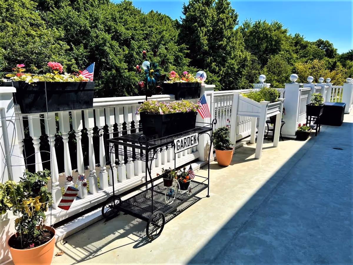 Outdoor patio area with white railing and various potted plants and flowers. Several American flags are placed among the plants. A black metal cart with the word 'GARDEN' on it holds flower boxes. Trees and blue sky are visible in the background.