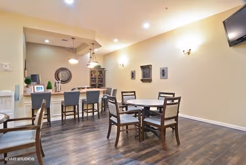 A well-lit dining area with a round table and four chairs in the foreground, and a counter with four high chairs in the background. The room has wooden flooring, beige walls with wall sconces, decorative wall art, and a mounted flat-screen TV in the upper right corner.