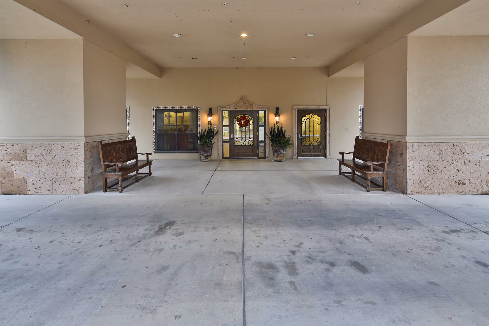 Covered entrance area of a building with two wooden benches on either side, two potted plants flanking a decorative glass door with a holiday wreath, and a smaller door to the right. The walls are beige with stone accents at the bottom.