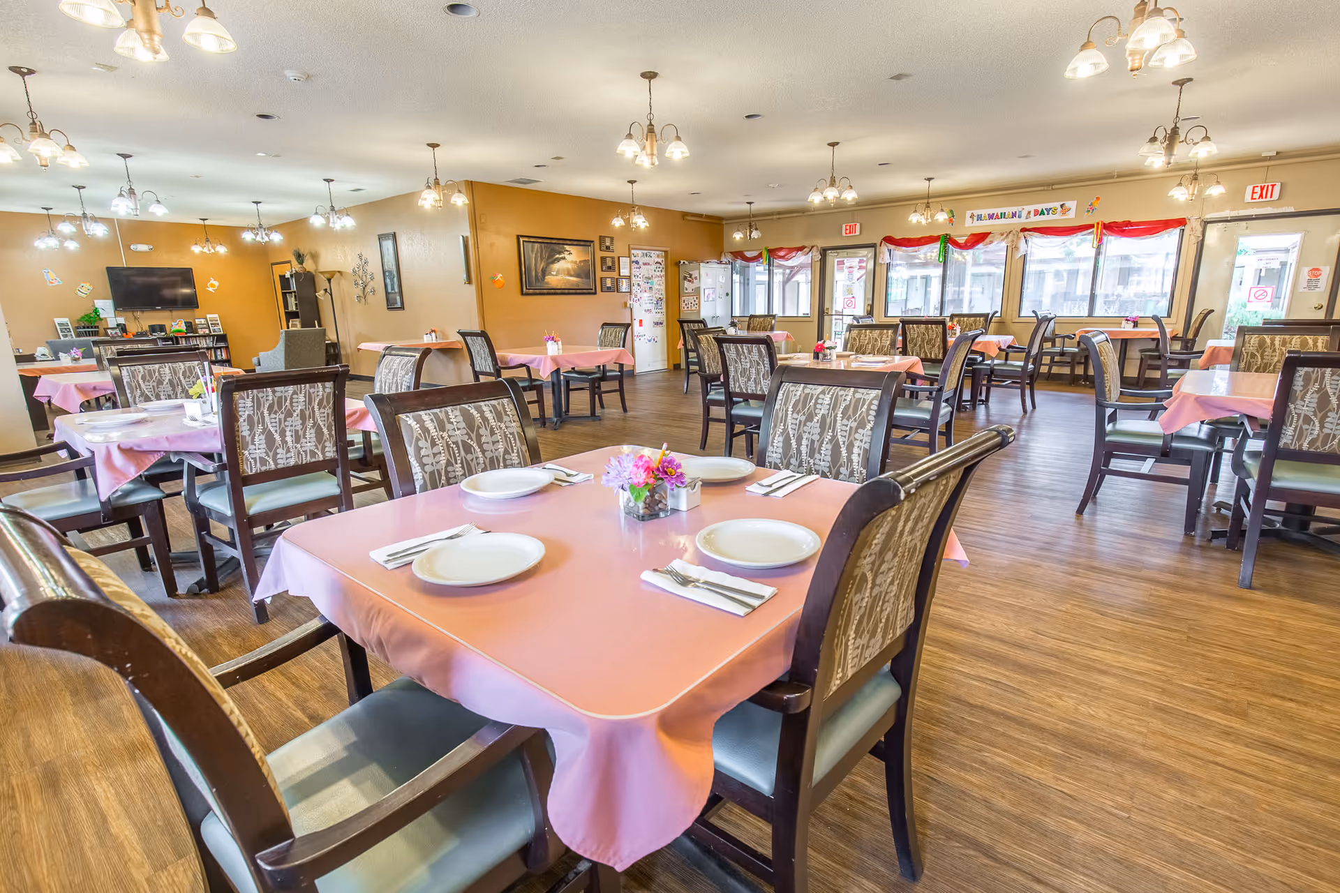 A spacious dining room with multiple tables covered in pink tablecloths, each set with white plates, napkins, and silverware. The room has wooden flooring, patterned cushioned chairs, and several ceiling light fixtures. Large windows line one side of the room, allowing natural light to fill the space. There is a television and bookshelf in the background, along with some wall decorations and exit signs above doors.
