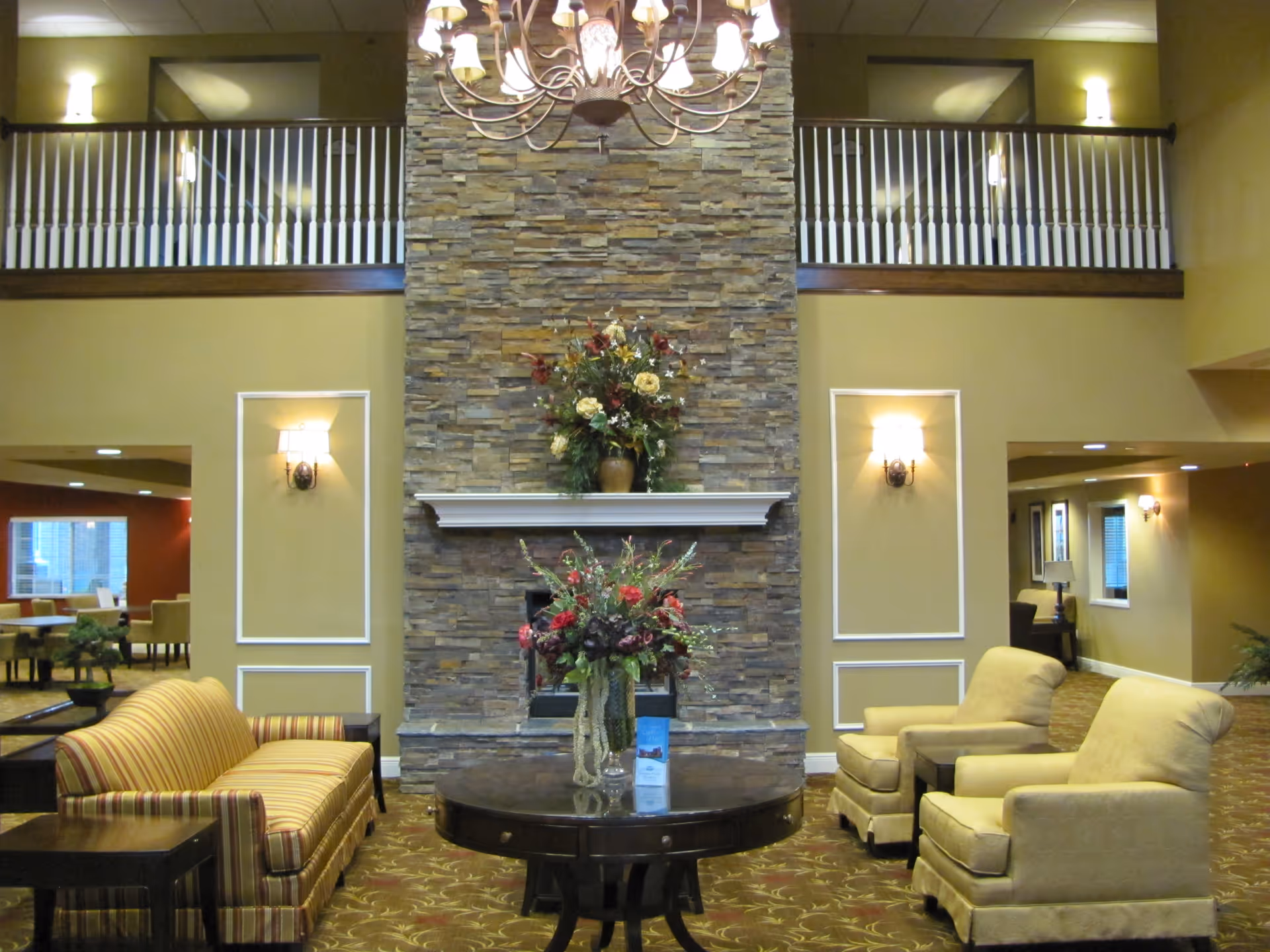 A spacious and warmly lit living room area in a senior living facility featuring a tall stone fireplace with a floral arrangement on the mantel and another on a round wooden table in front. The room has a striped sofa on the left, two beige armchairs on the right, wall sconces, and a chandelier hanging from the ceiling. There is a balcony railing visible above the fireplace and additional seating areas in the background.