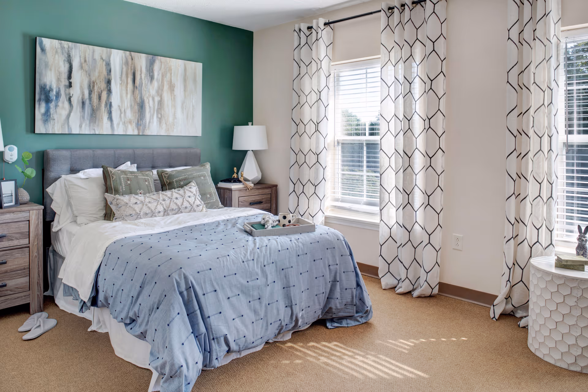 A cozy bedroom with a neatly made bed featuring a blue patterned comforter and multiple pillows. The bed has a gray upholstered headboard and is flanked by two wooden nightstands, one with a white lamp and the other with decorative items. Behind the bed is a green accent wall with an abstract painting. Two windows with white and black patterned curtains allow natural light to fill the room. The floor is carpeted in a light brown color.