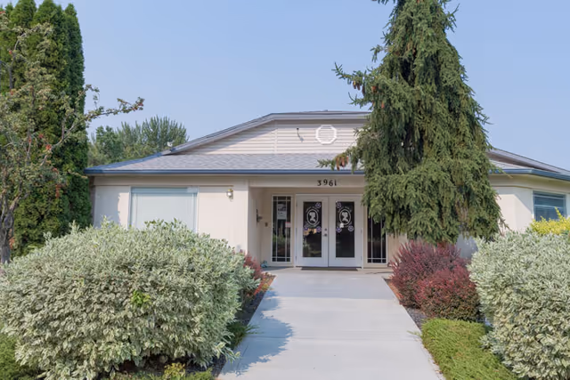 Front exterior view of Ashley Manor Memory Care building with a concrete walkway leading to double glass doors. The building is surrounded by green shrubs, bushes, and tall trees under a clear sky.