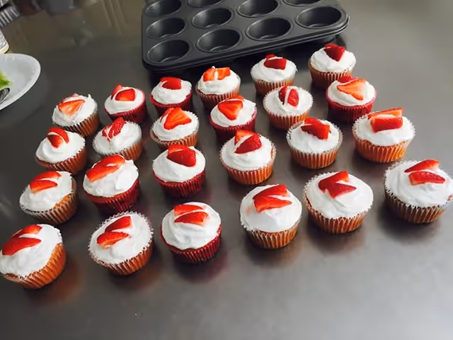 A group of cupcakes with white frosting and sliced strawberries on top arranged on a metal countertop next to a black muffin tray.