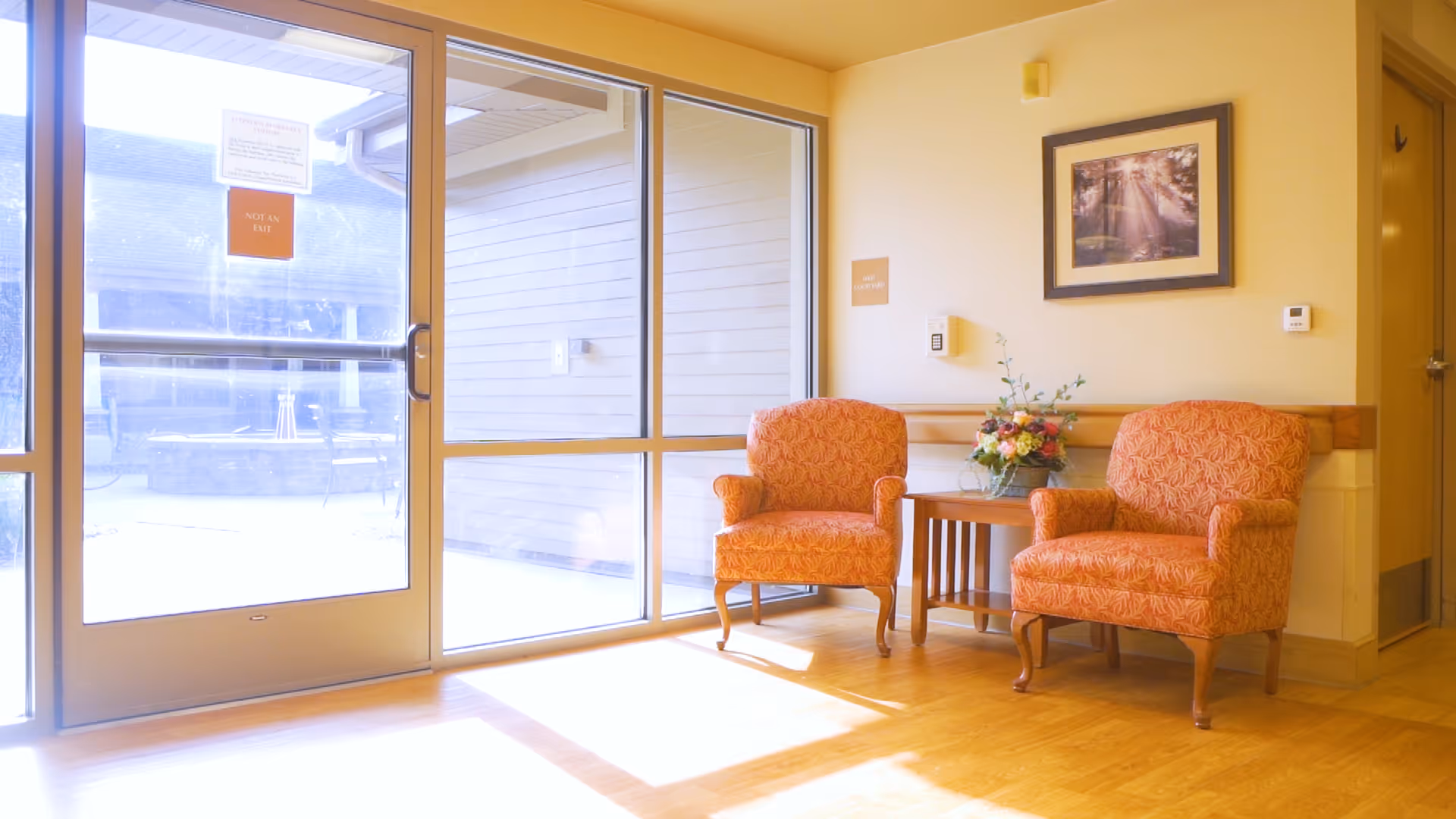 A bright seating area in a senior living facility with two orange patterned armchairs and a small wooden table with a floral arrangement between them. There is a large glass door and windows letting in sunlight, and a framed picture on the wall above the chairs.