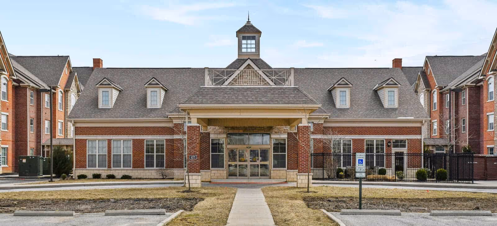 Front exterior view of a large brick building with multiple windows and a central entrance covered by a small roof. The building has a gray shingled roof with dormer windows and a small tower-like structure on top. There is a paved walkway leading to the entrance and a parking area with handicap parking signs in front.