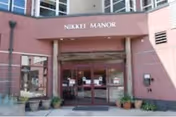 Entrance of a building with a sign above the door that reads 'Nikkei Manor'. The entrance has glass double doors and several potted plants placed around the doorway.
