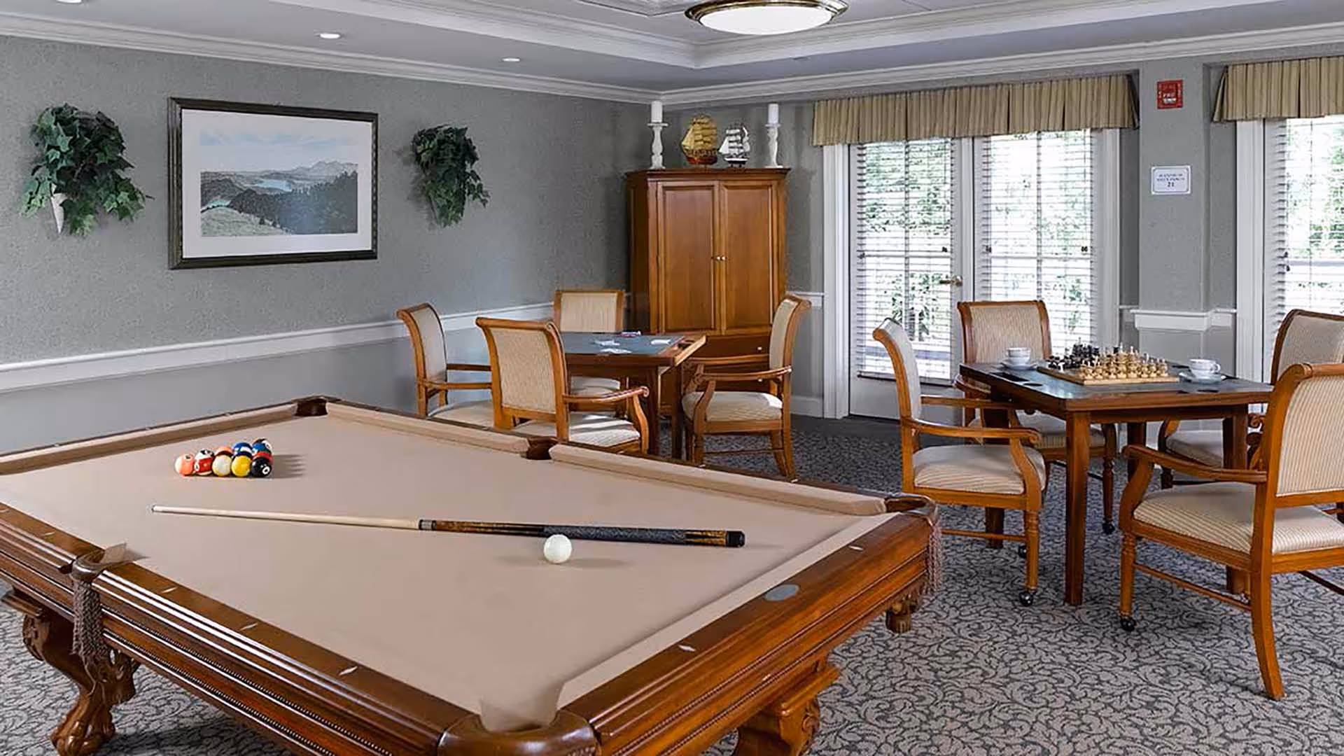 A recreational room featuring a pool table with a cue stick and billiard balls arranged for a game. In the background, there are two tables with chairs, one set up for a card game and the other with a chessboard and cups. The room has large windows with blinds, a wooden cabinet, and decorative plants on the walls.