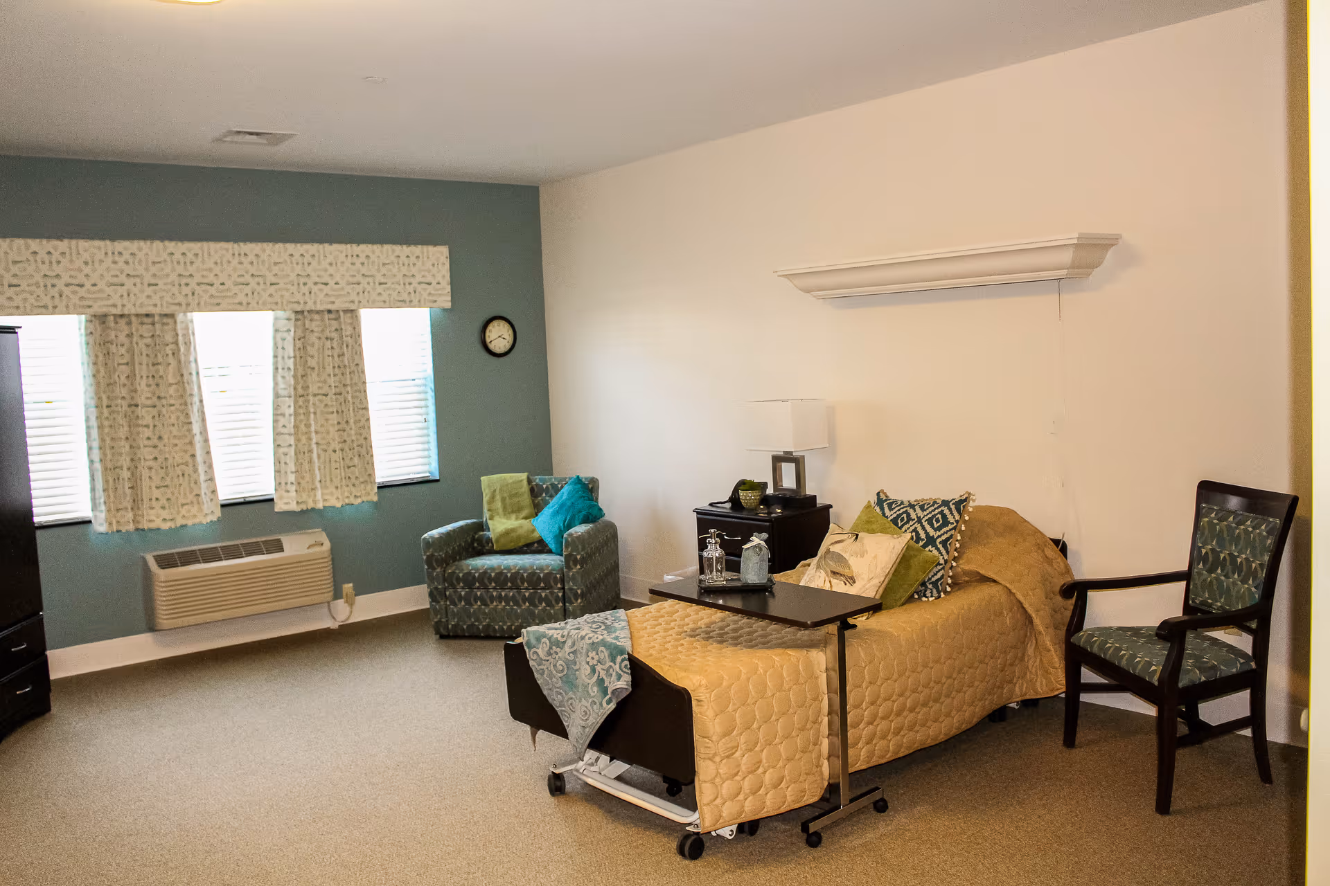 A furnished resident bedroom with a single hospital-style bed, armchair, side table, and window with curtains.